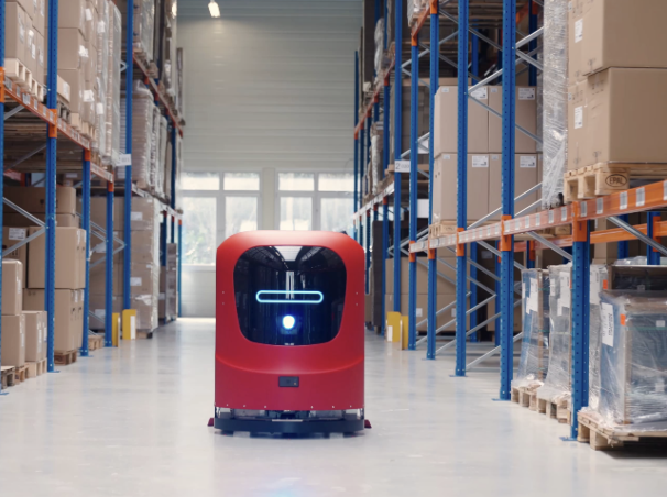 Red autonomous floor-cleaning robot in a warehouse aisle between storage racks